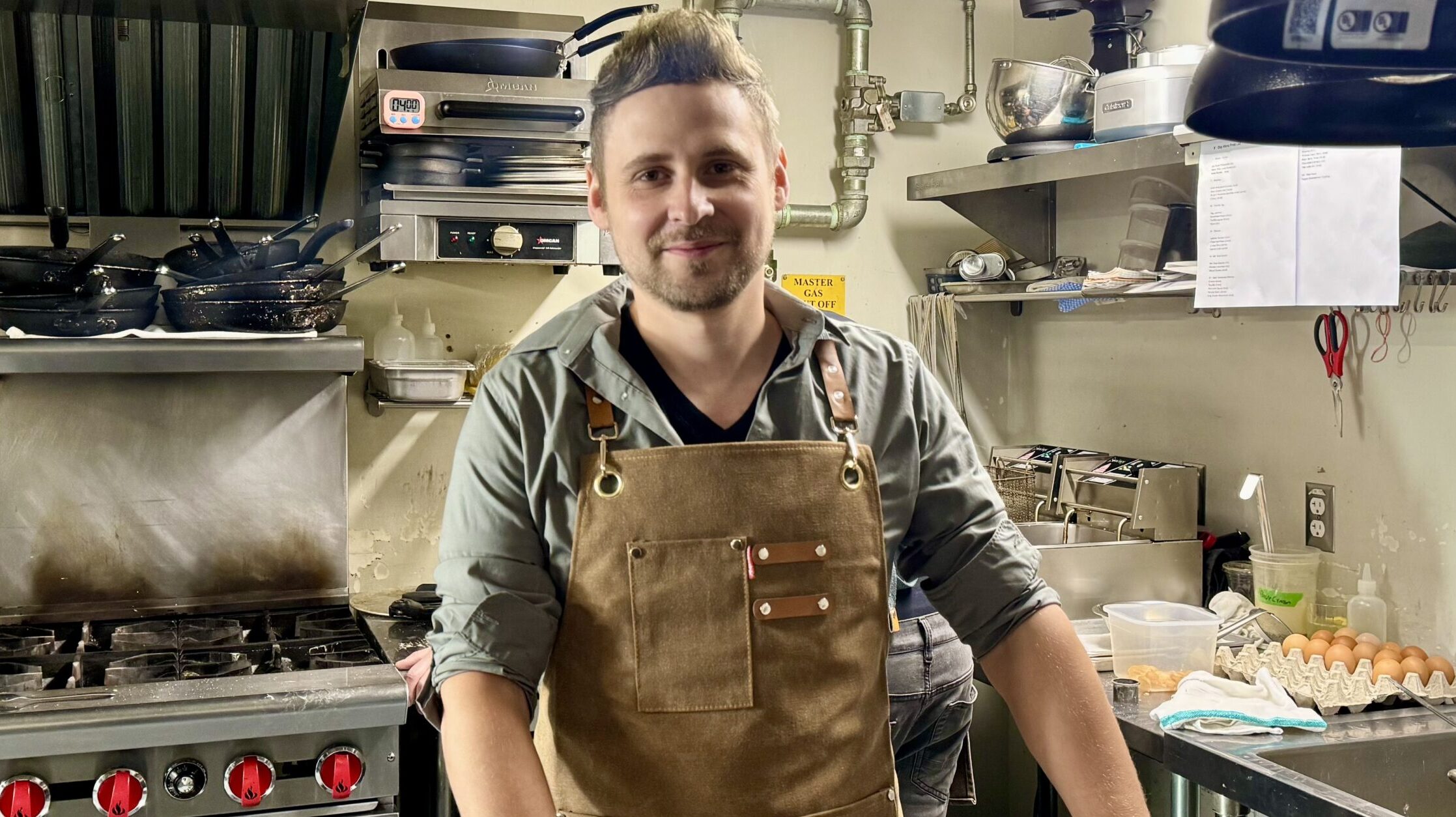 Man in a green shirt and beige apron stands in a kitchen smiling.