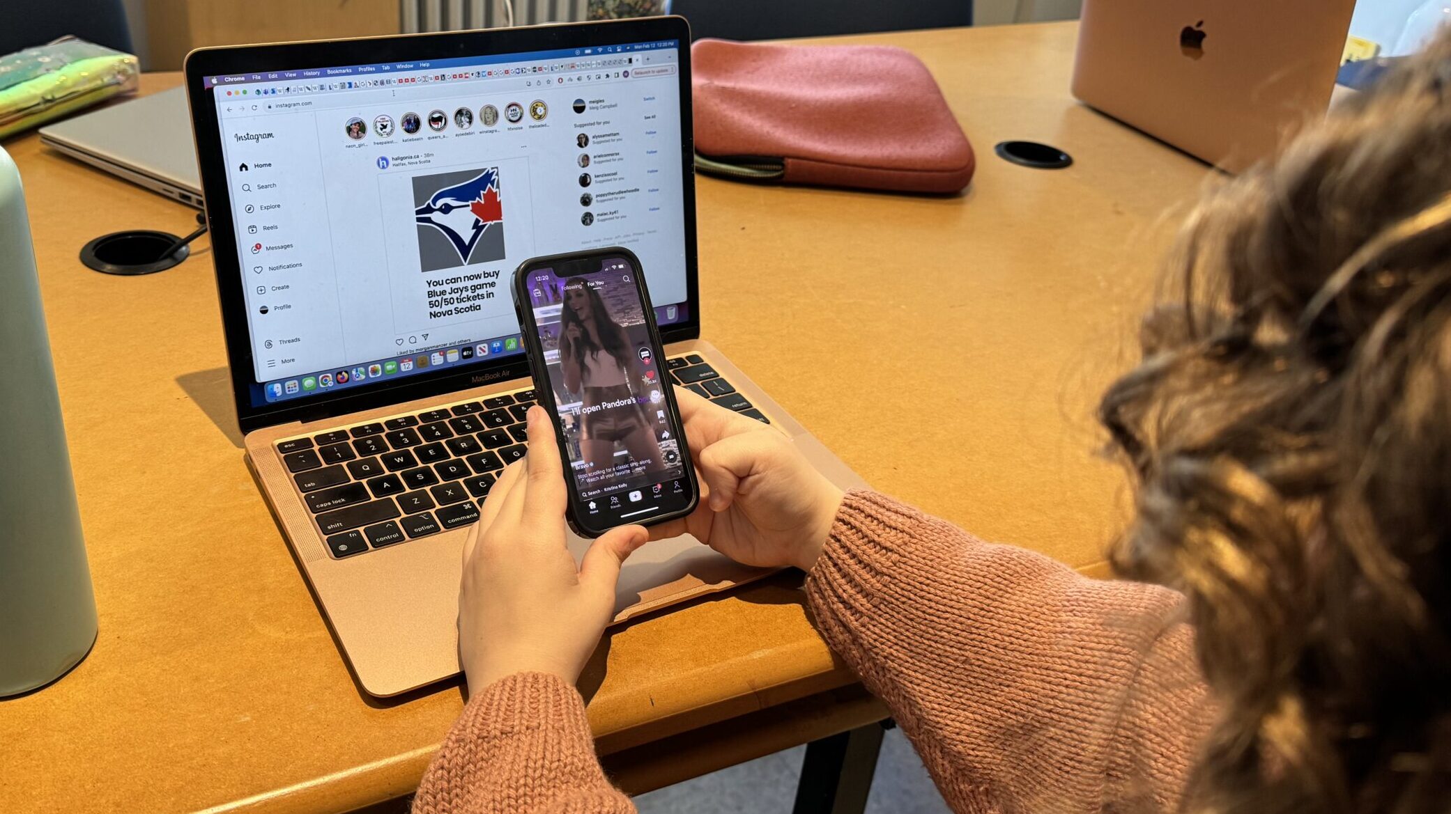 Woman in a pink cardigan uses her smartphone to use TikTok and her laptop for Instagram.