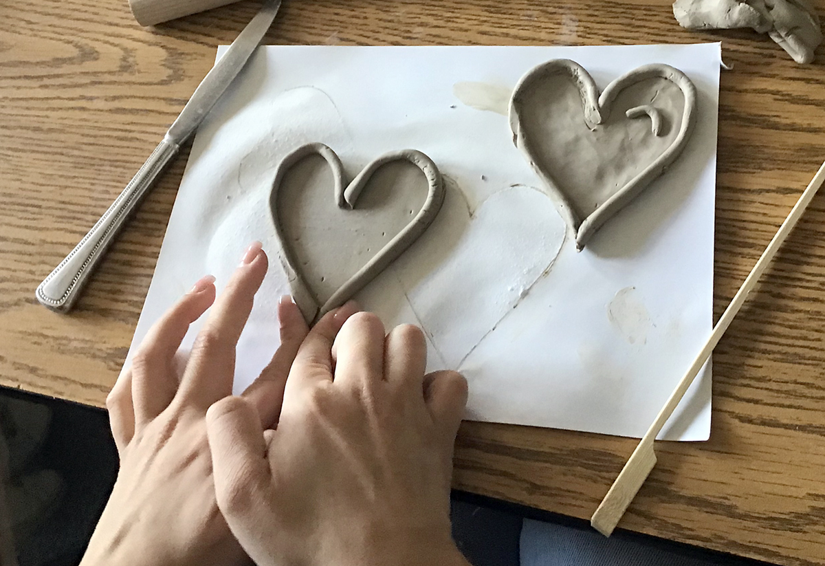 Two hands form a heart-shaped piece of clay on a wooden table