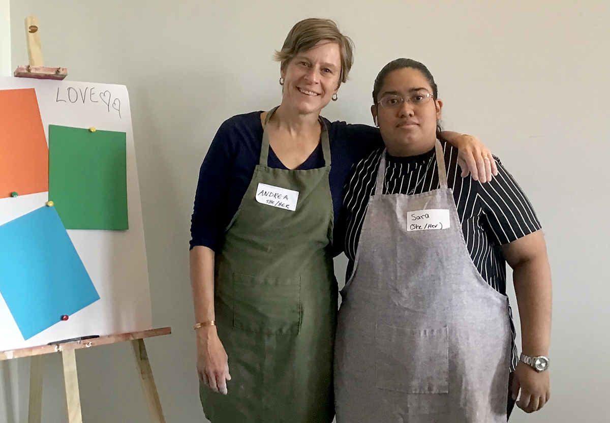 Two people in wearing artist aprons stand beside an easel