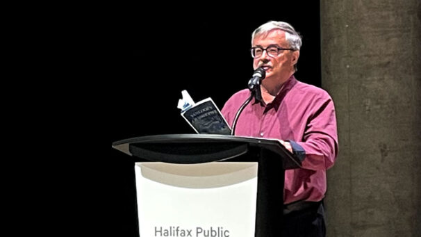 A man in a red shirt stands at a podium while holding a book.