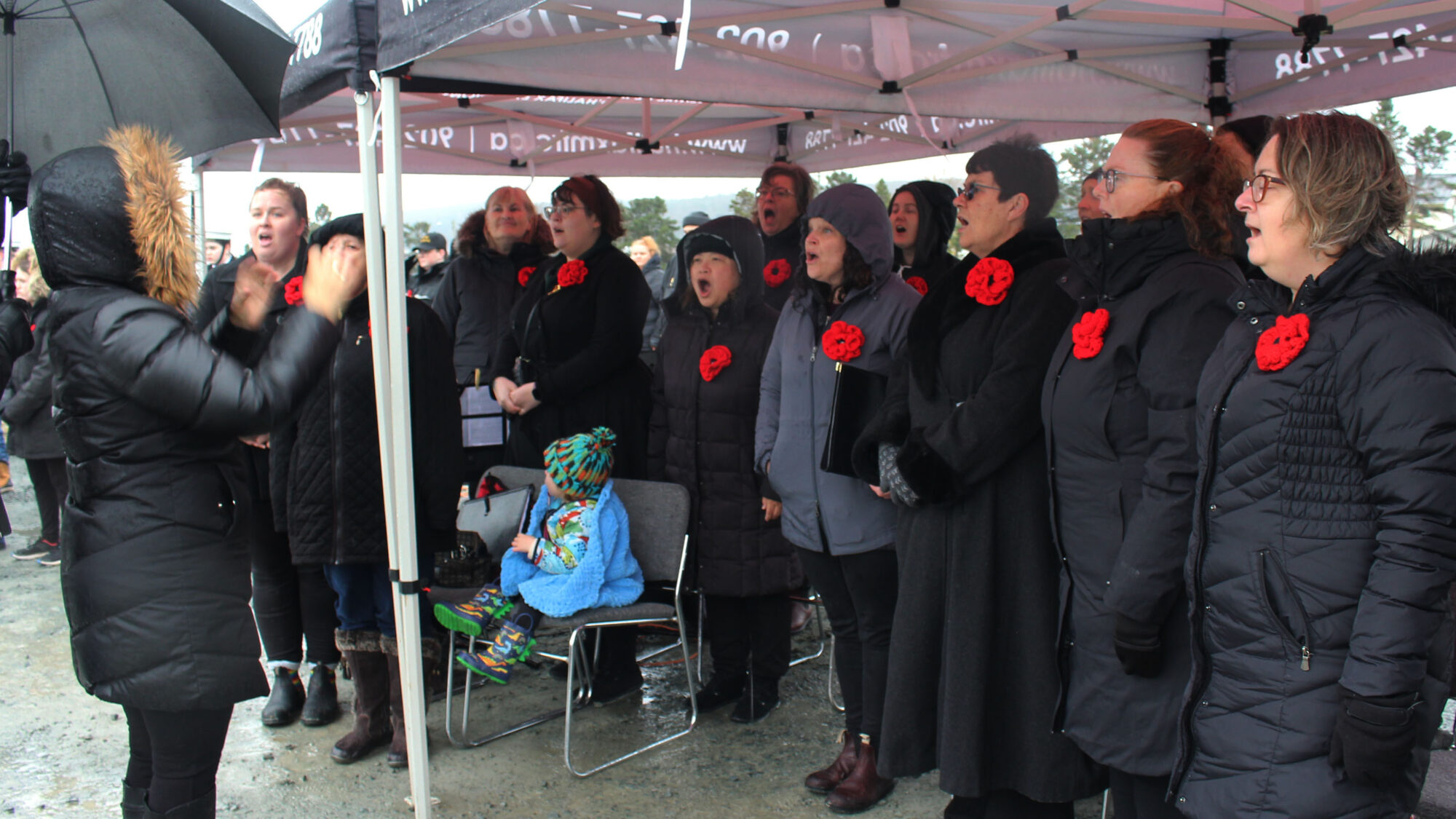 Women with red poppies pinned to their black jackets sing while a woman conducts.