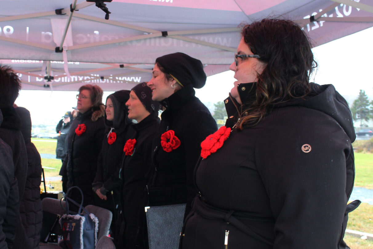 Women stand in a line and sing with red poppies pinned to their black jackets.