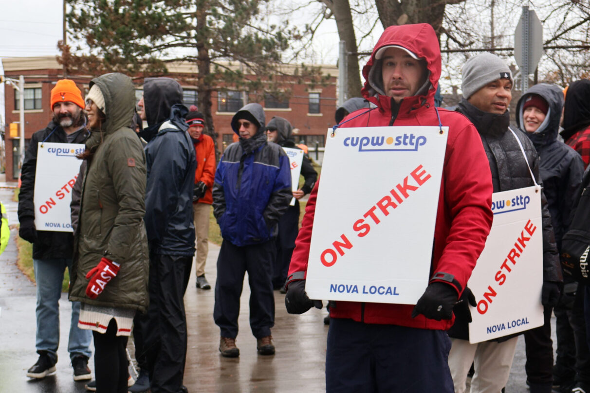 A man in a red jacket has a sign reading "on strike" hanging from around his neck.