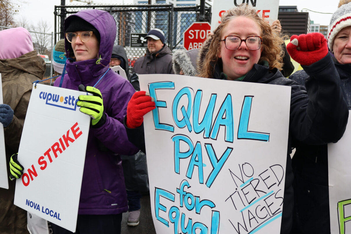 Two female postal workers hold signs reading "on strike" and "equal pay for equal work."