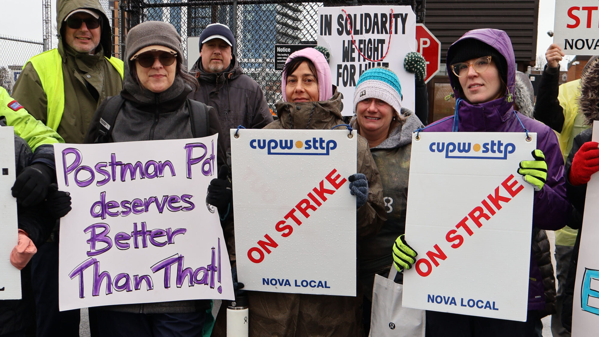 Three women hold signs on the picket line. One sign reads "Postman Pat deserves better than that!" The other two read "on strike."