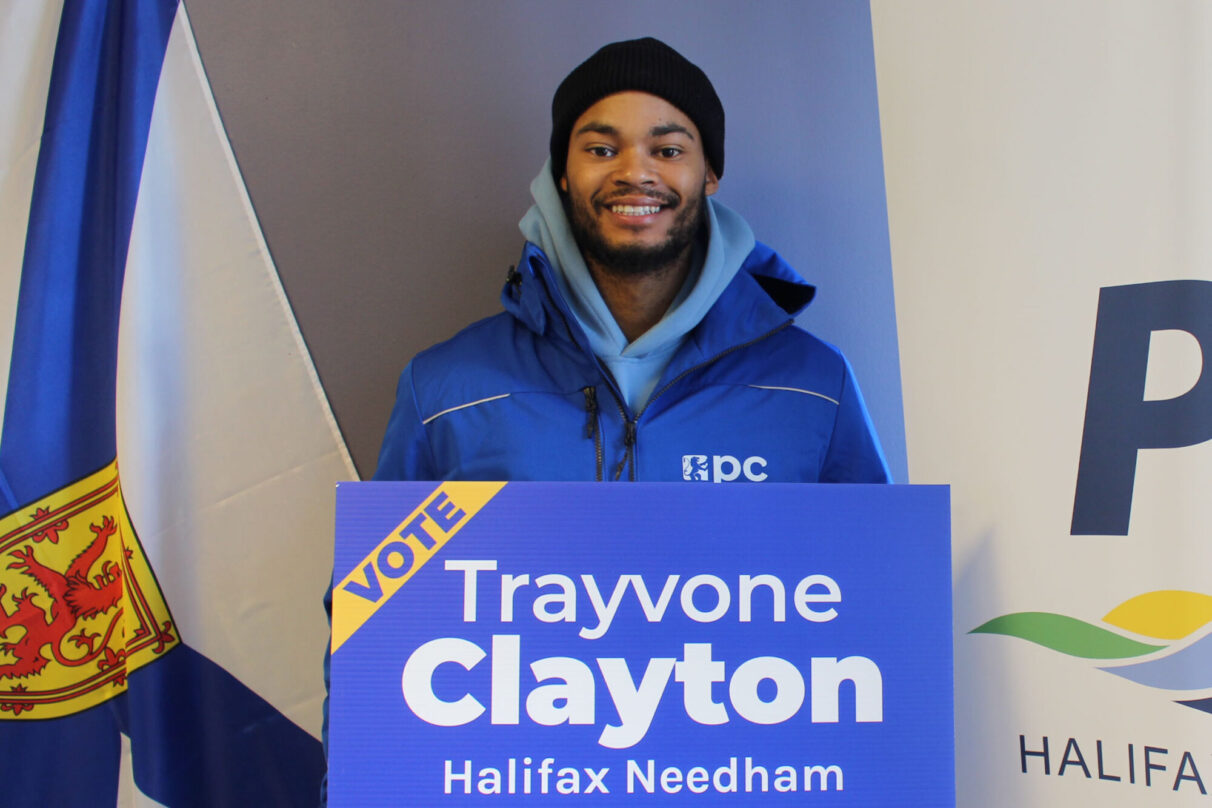 A man stands with a sign reading "Vote Trayvone Clayton Halifax Needham"