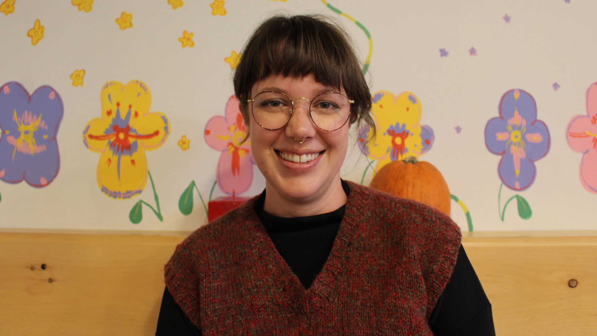 A woman in a red sweater vest sits in front of a floral background. She smiles at the camera.