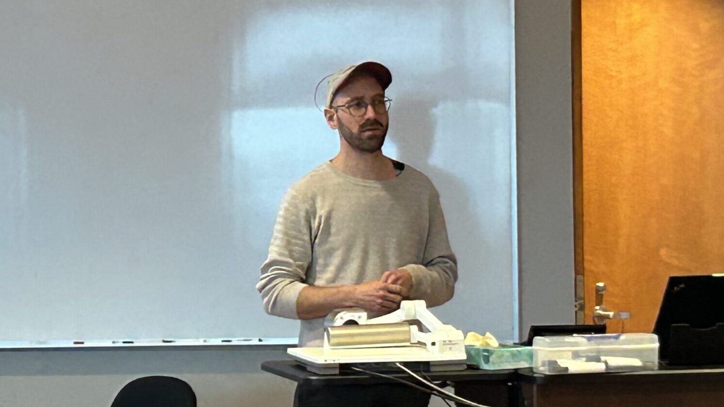 A man in a ball cap delivers a lecture in front of a white board.