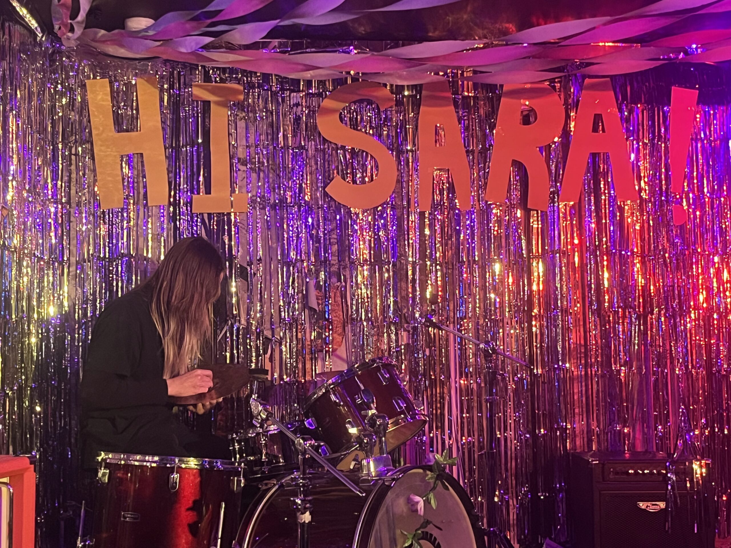 A girl sits at a drumkit. The walls of the stage are lined with tinsel. Paper letters read "HI SARA!" above her head.