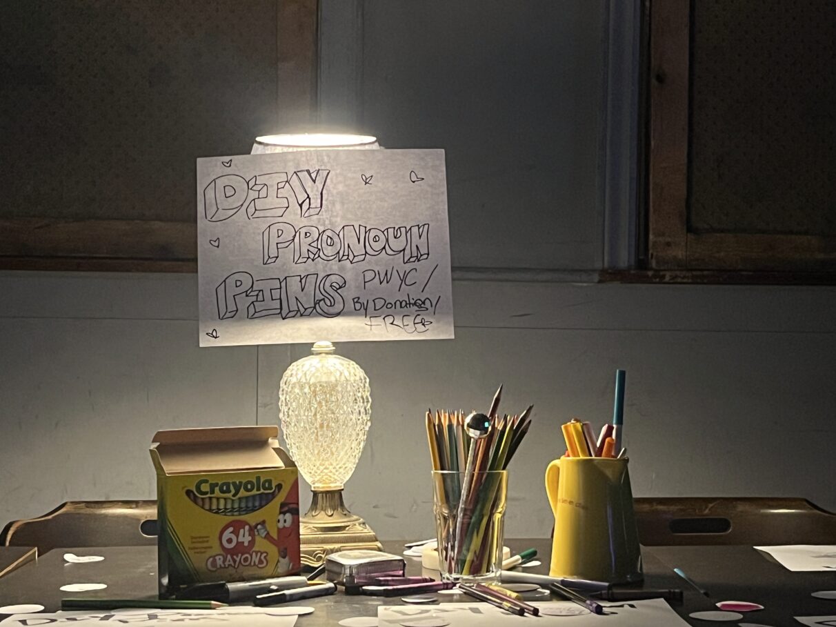 Sign that reads "DIY Pronoun Pins PWYC/By Donation/Free" is taped to a lamp. Crayons and pencils are next to it.