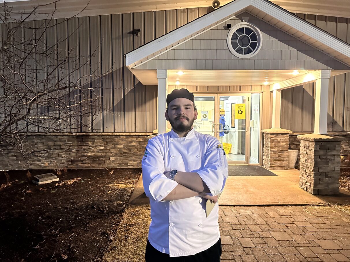 A man standing outside a polling station with arms folded wearing a chef's uniform.