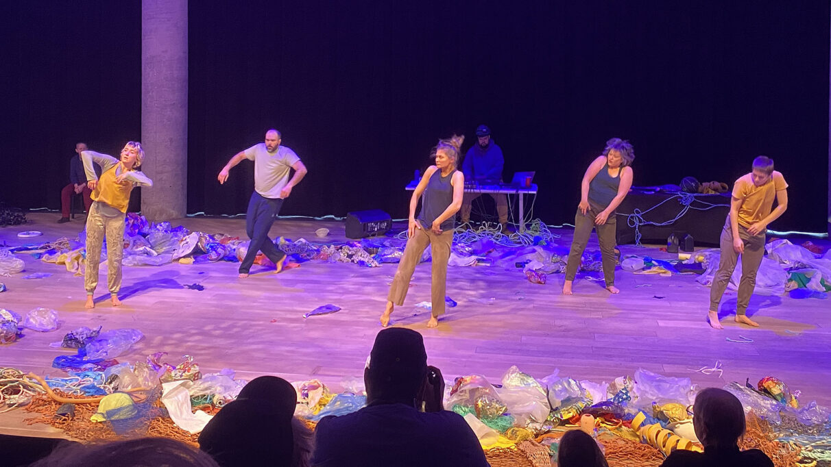 5 dancers under purple stage lighting surrounded by plastic waste