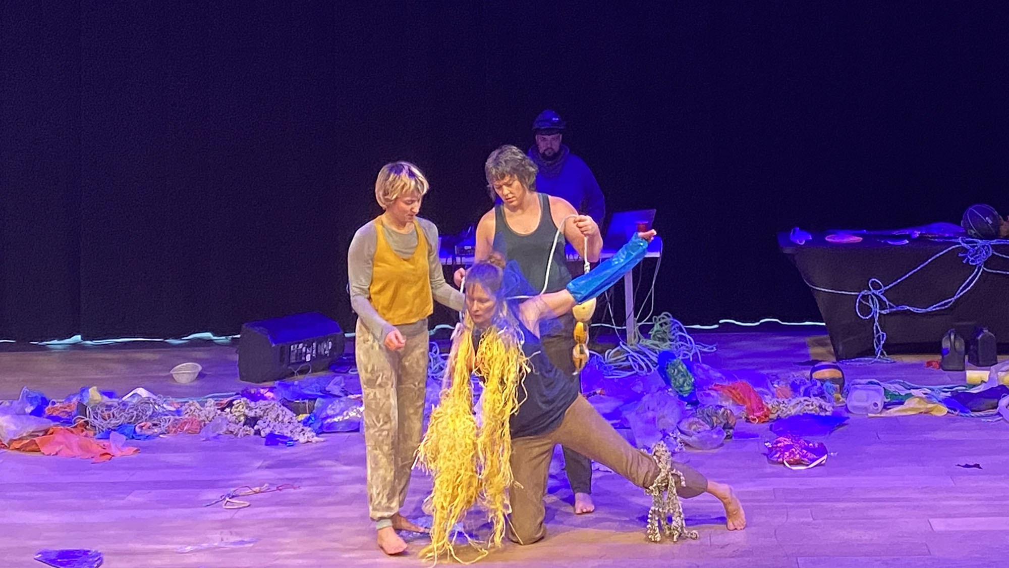 Three dancers with piled plastic waste behind them, one dancer has yellow plastic hanging around her neck