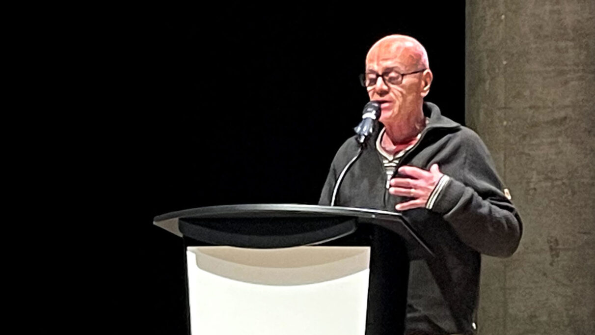 A man in a black sweater reads a poem at a podium.