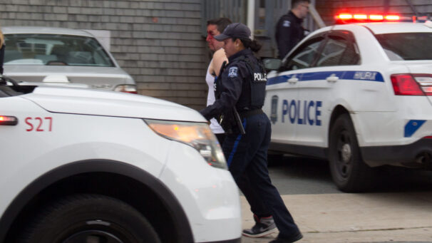 A man wearing a white tank top, with a face covered in blood, is escorted to a police vehicle by an officer.