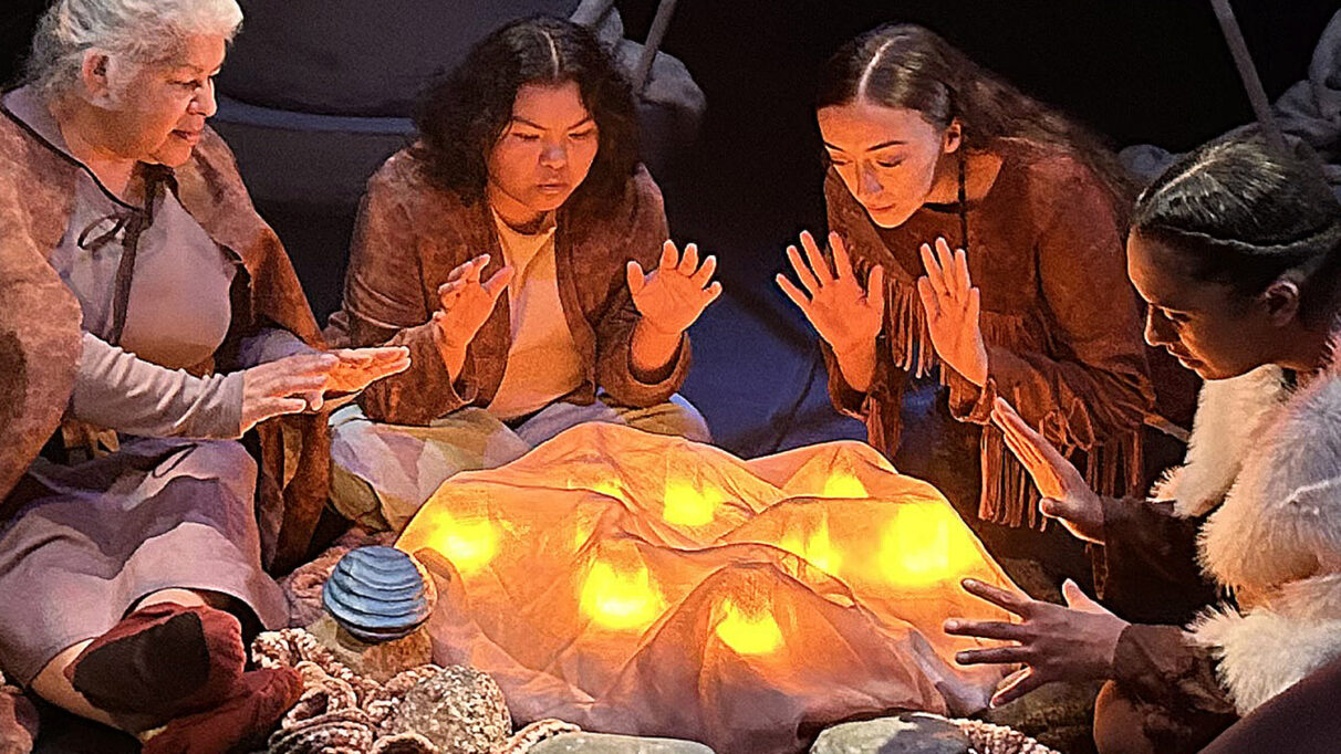 Four performers heat themselves around a fire