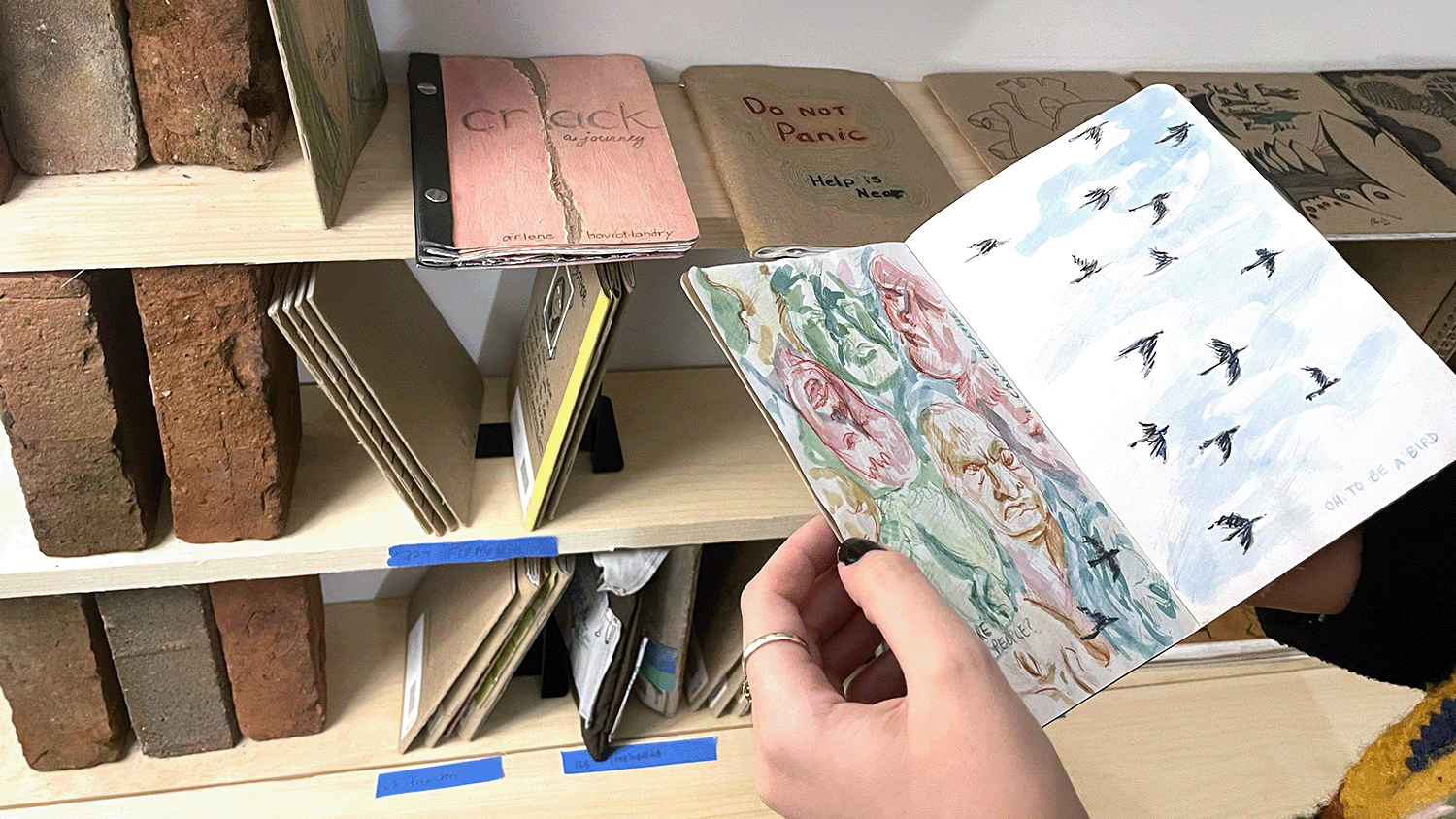 Hands holding a sketchbook in front of shelves filled with other similar sketchbooks.