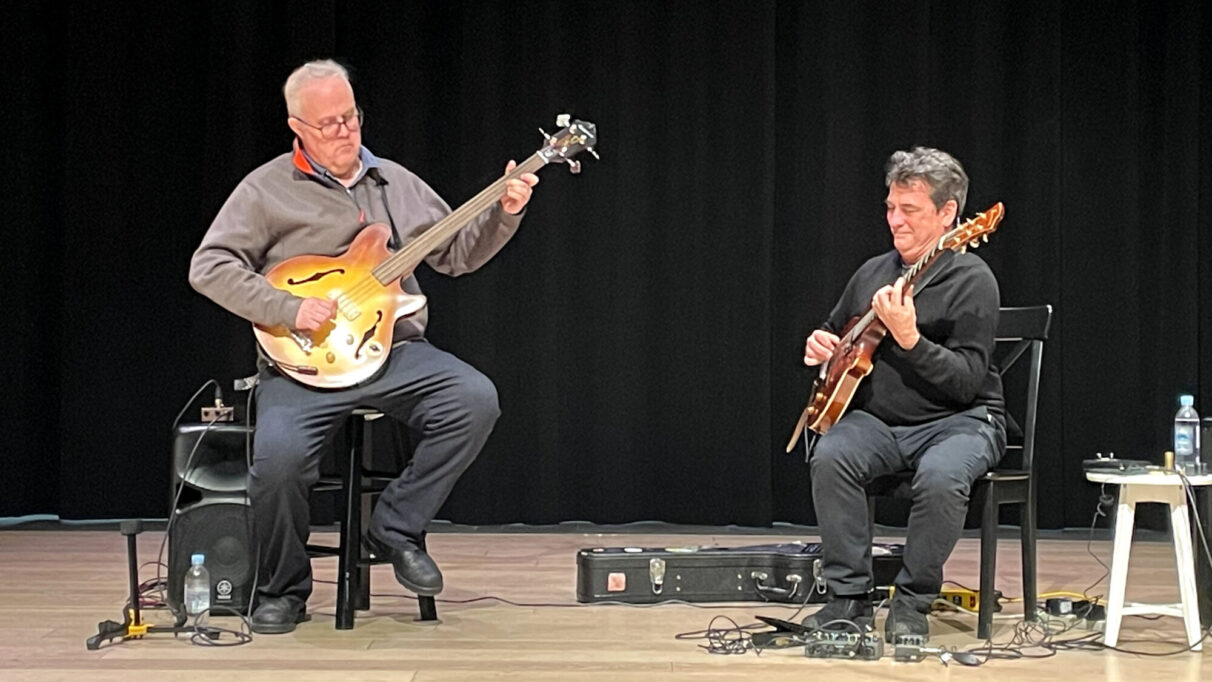 Two men play guitars sitting next to each other.