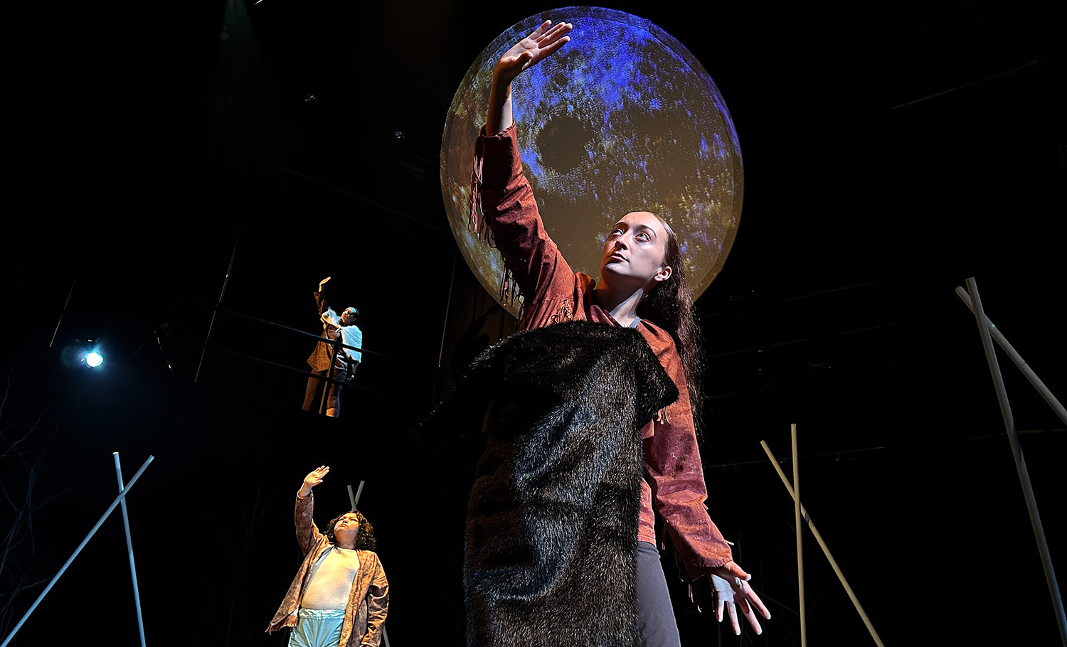 Three dancers reach to the sky in front of a moon projection