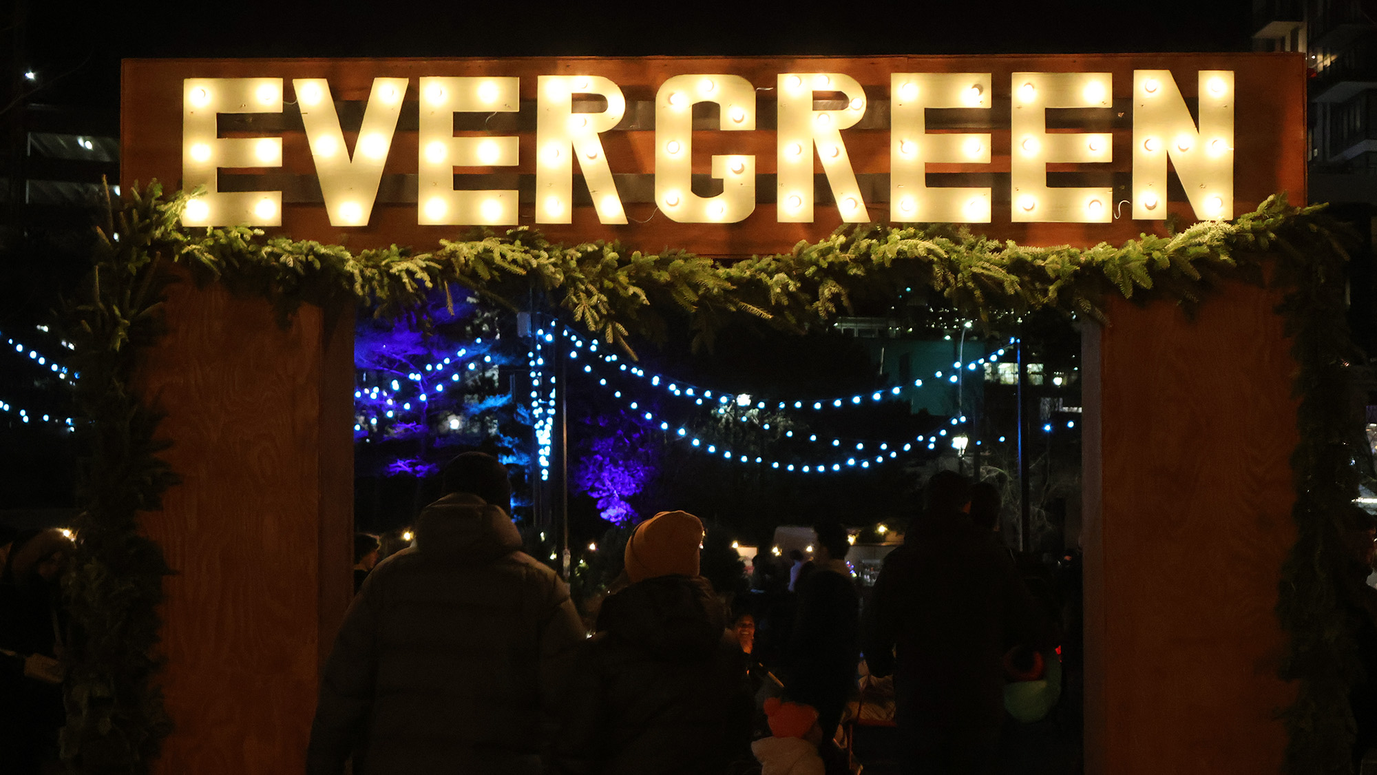 The word 'evergreen' is lit up above a wooden archway with people walking underneath. There are strings of lights in the background.