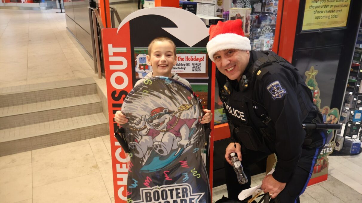 A young boy holds up a new sled with a polar bear on it while standing next to a police man in a Santa hat.