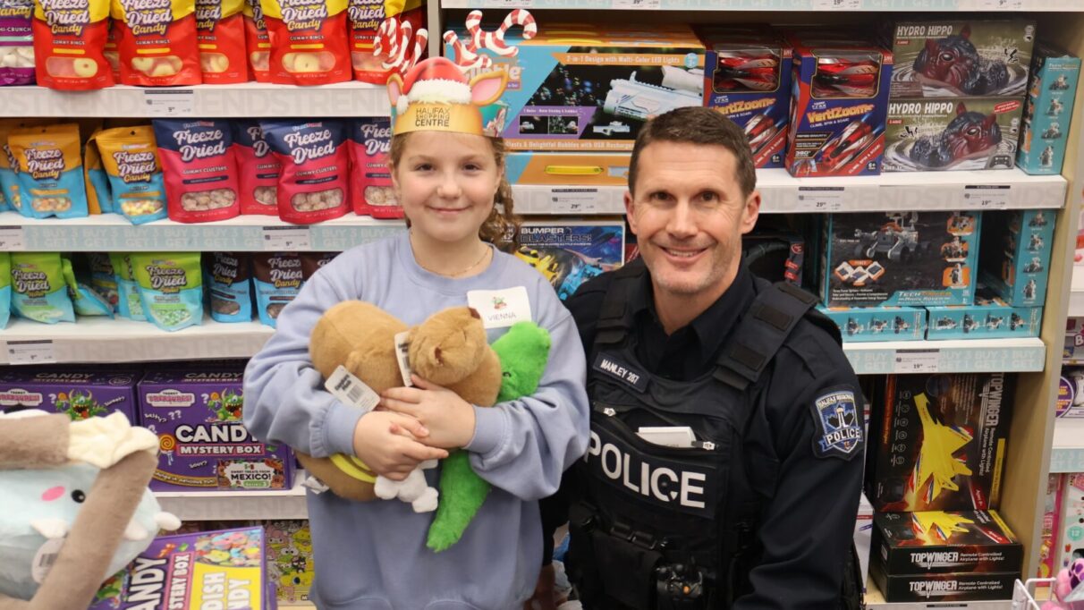 A police officer and a kid wearing reindeer antlers poses in front of a shelf filled with candy and toys.