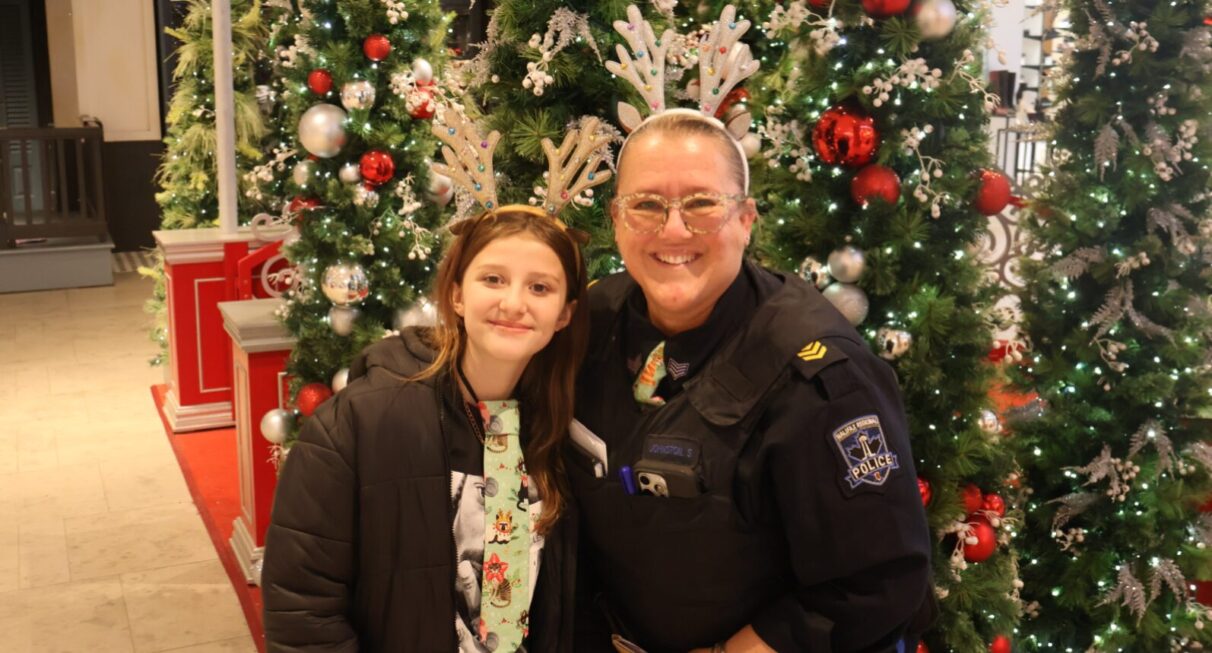 A police officer wearing reindeer antlers poses alongside a young girl also wearing reindeer antlers in front of a collection of Christmas trees