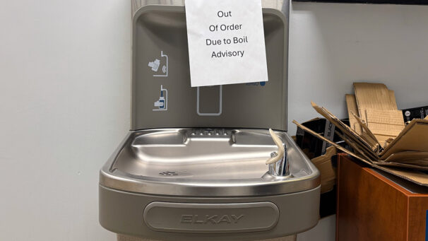 A sign taped to a water fountain says "Out Of Order Due to Boil Advisory."