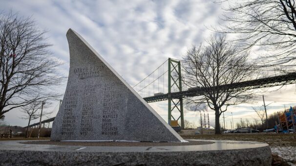 a grey stone memorial carved with last names. A green toll bridge looms in the background under a cloudy sky.