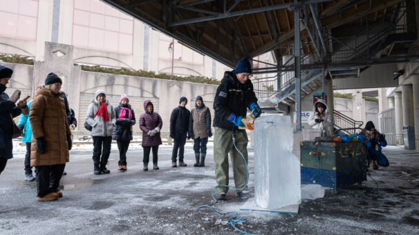 a man takes a chain saw top a block of ice while he is surround by people watching him work.