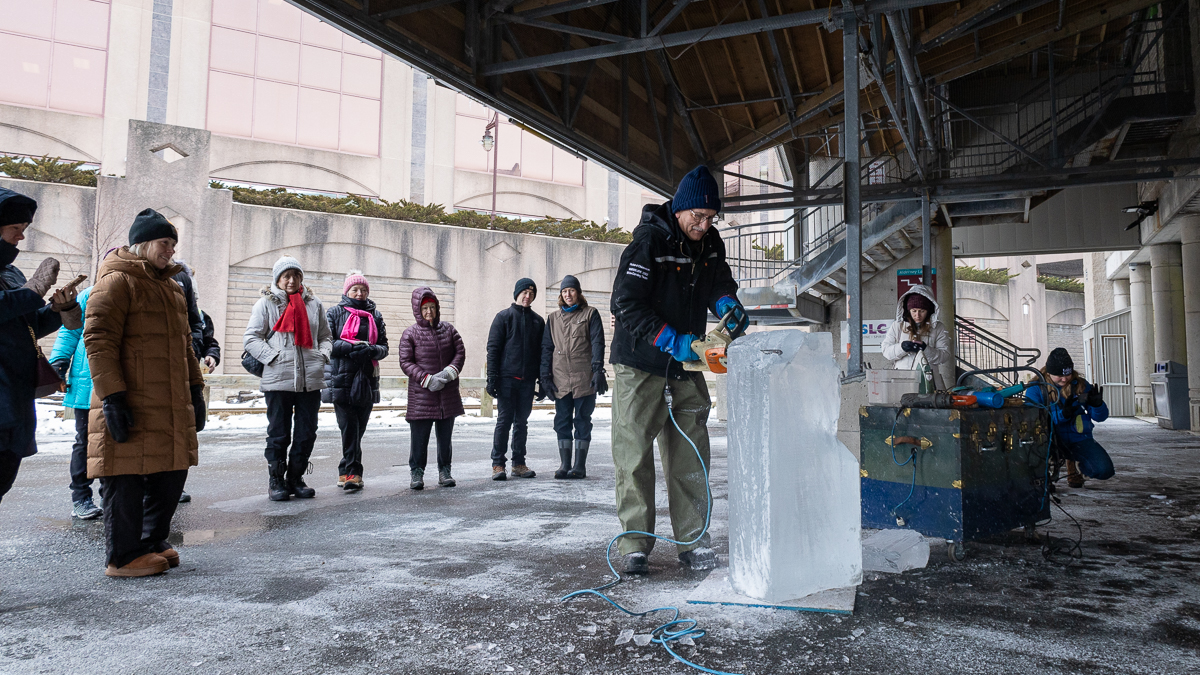 a man takes a chain saw top a block of ice while he is surround by people watching him work.