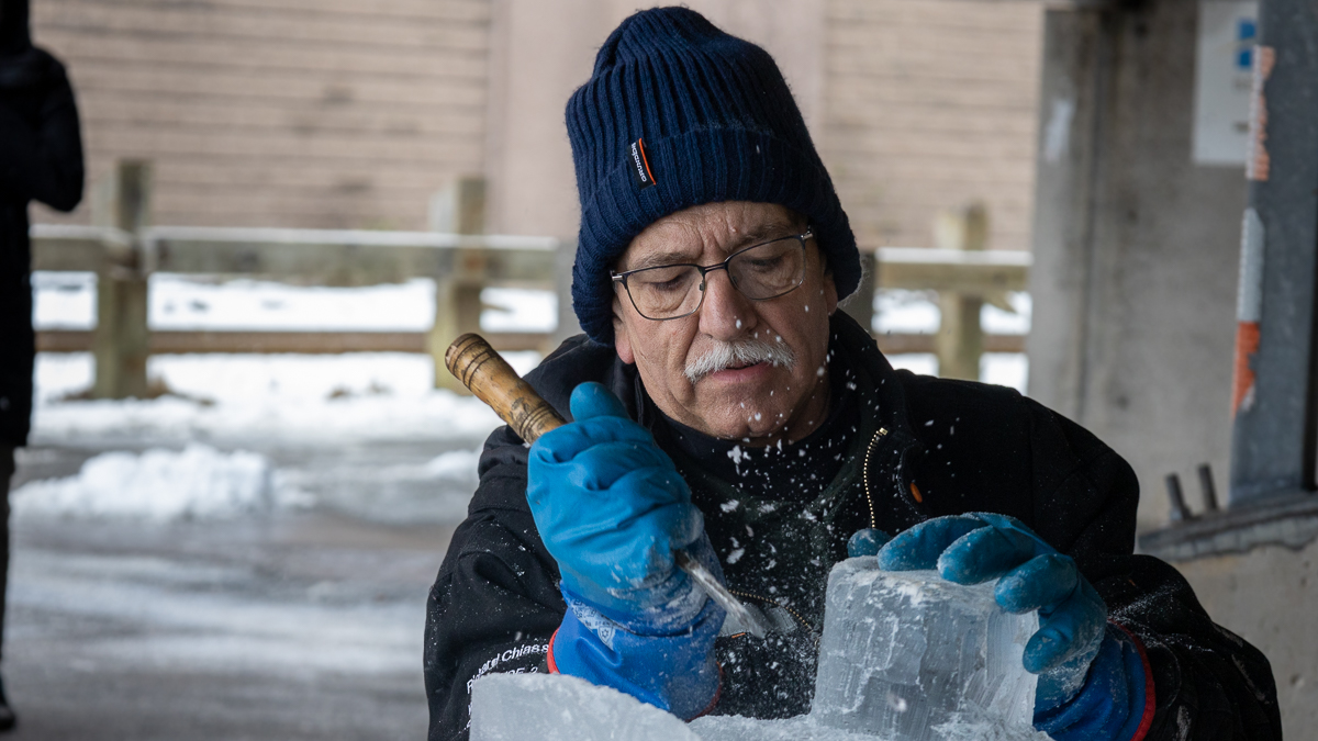 a man wearing a blue hat and glasses chips away at an ice block using a metal tool, he is wearing blue gloves.