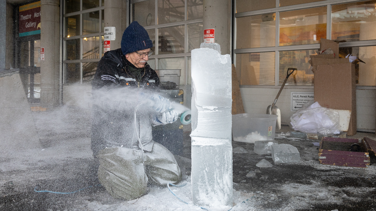 a man rests on his knees, shaving away ice on a large block using a power tool.