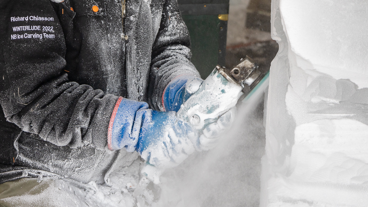 A close up of a blue pair of gloves grinding away the ice as a man sculpts a snowman out of an ice block.