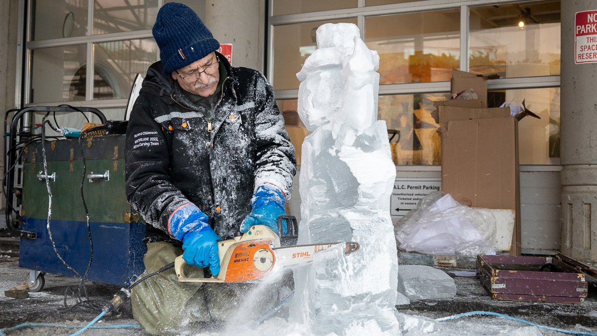 A na wearing a blue hat, black jacket, and green pants kneels in front of a block of ice and carves away at it using an orange and white chain saw.