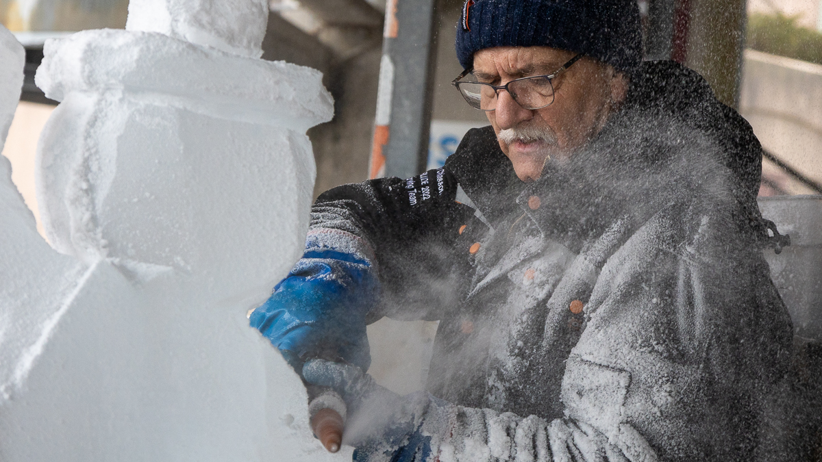 A man drills into a block of ice creating a snowman. He is wearing a black jacket and blue gloves.