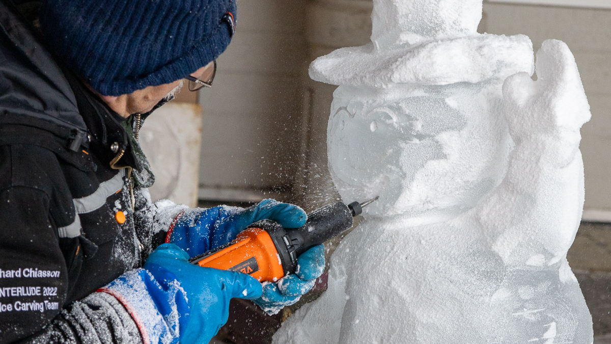 A man carves a smile into a snowman made of ice with a power tool.