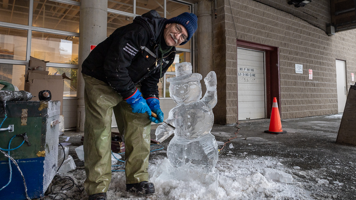 A man in a blue hat, black jacket, and green pants holds a metal tool used to carve ice. He stands beside a snowman made of ice.