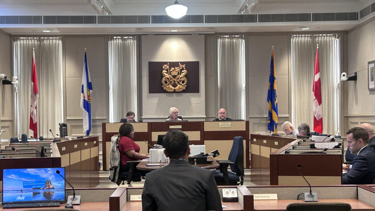 A city council meeting in session, with officials seated at a central podium and attendees at desks. Mervin David is back on facing the council.