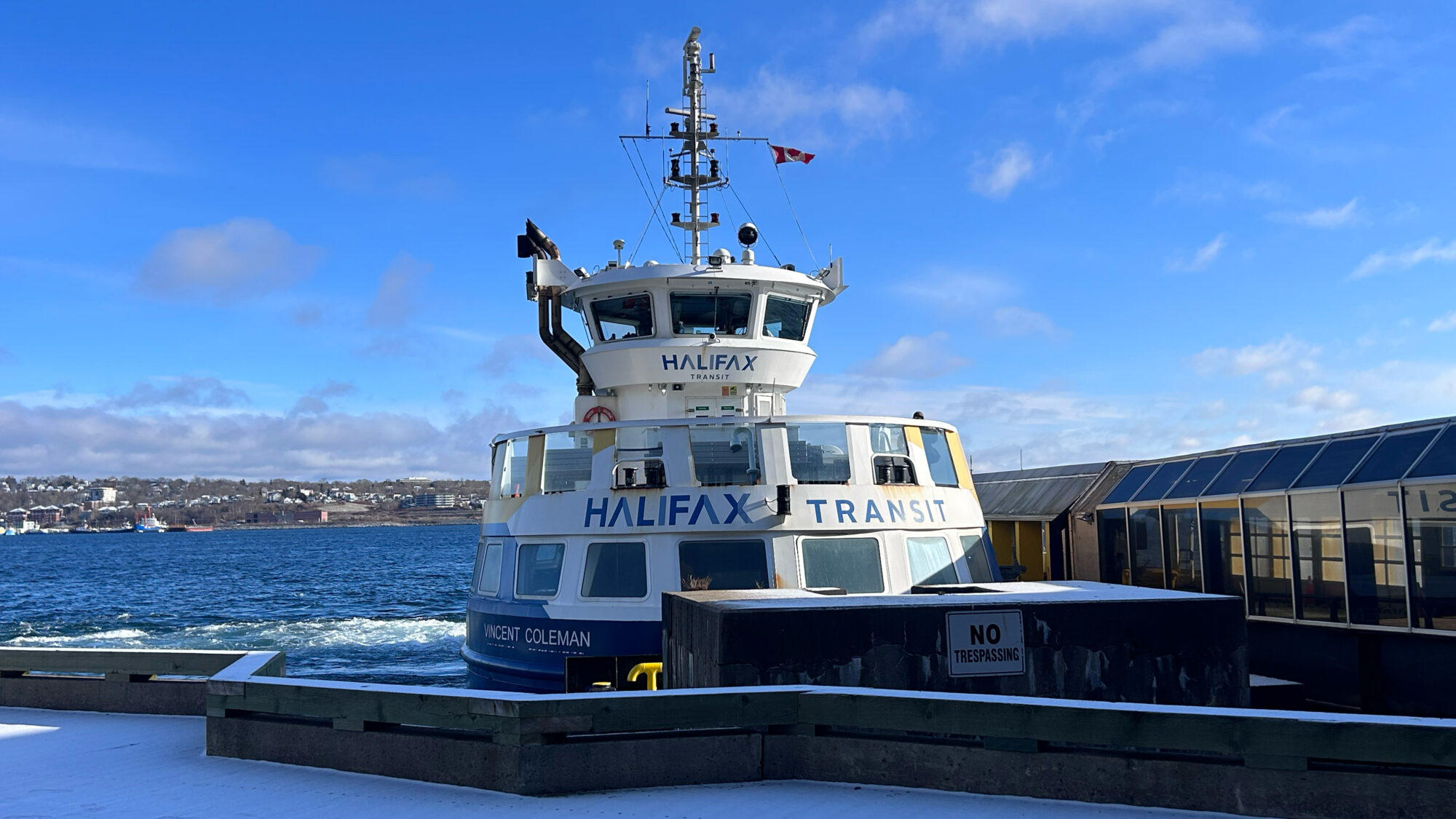 A Halifax Transit ferry at the dock on a sunny day, with cityscape and water in the background.