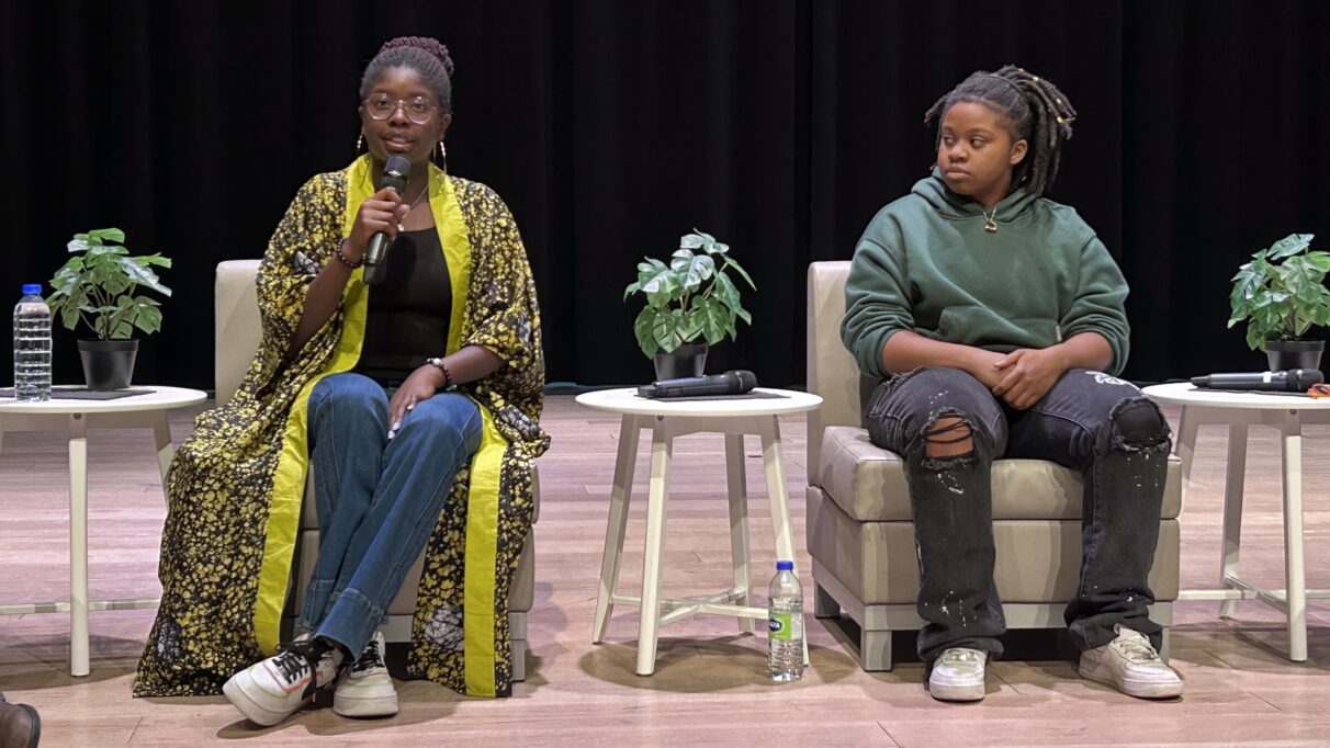 Two women sit next to each other on chairs with a table between them. The on the left is speaking into a microphone.