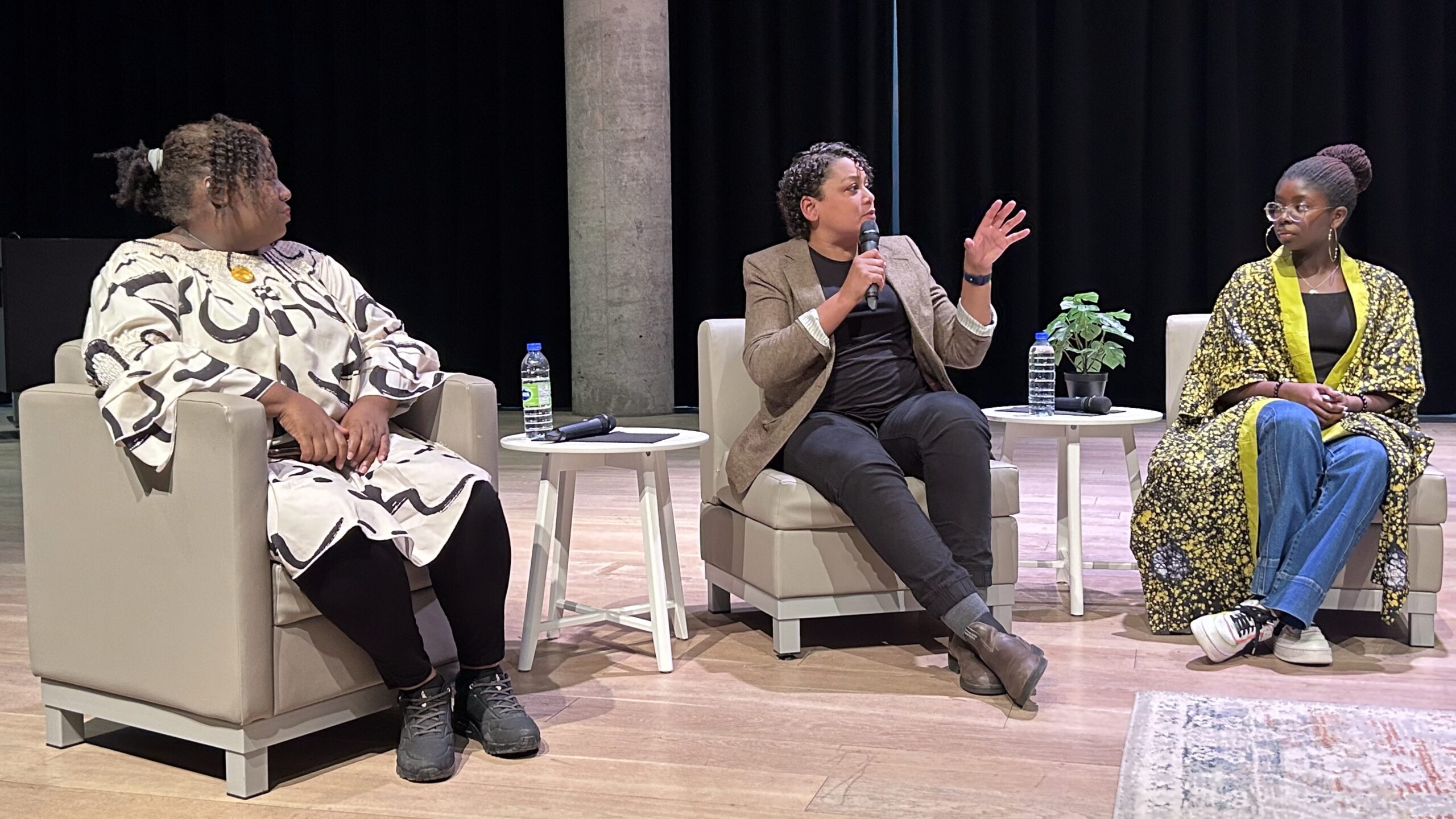 Thee women sit next to each other on chairs.The woman in the middle is speaking into a microphone.