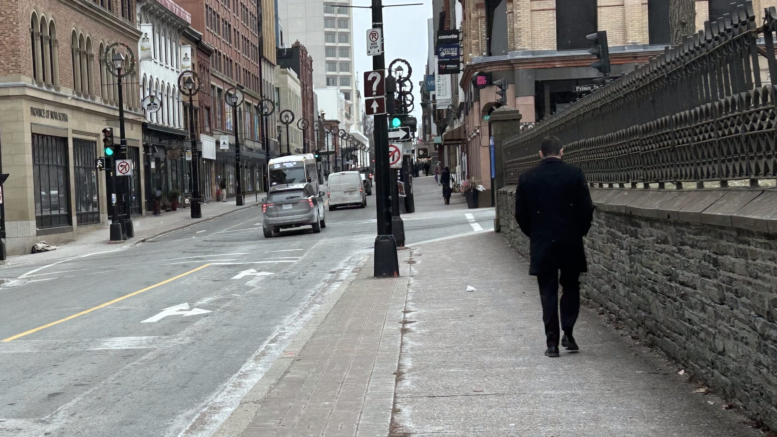 A man in a black coat walks on a city sidewalk next to a road with traffic and buildings lining the street.