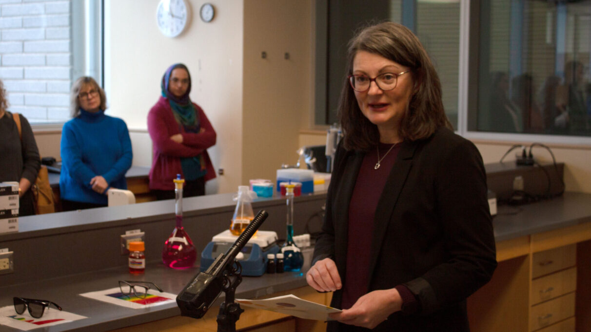 A woman in a black blazer and plum blouse speaks into a microphone. In the background are pink, yellow and blue beakers, and two people in a blue and a pink sweater.