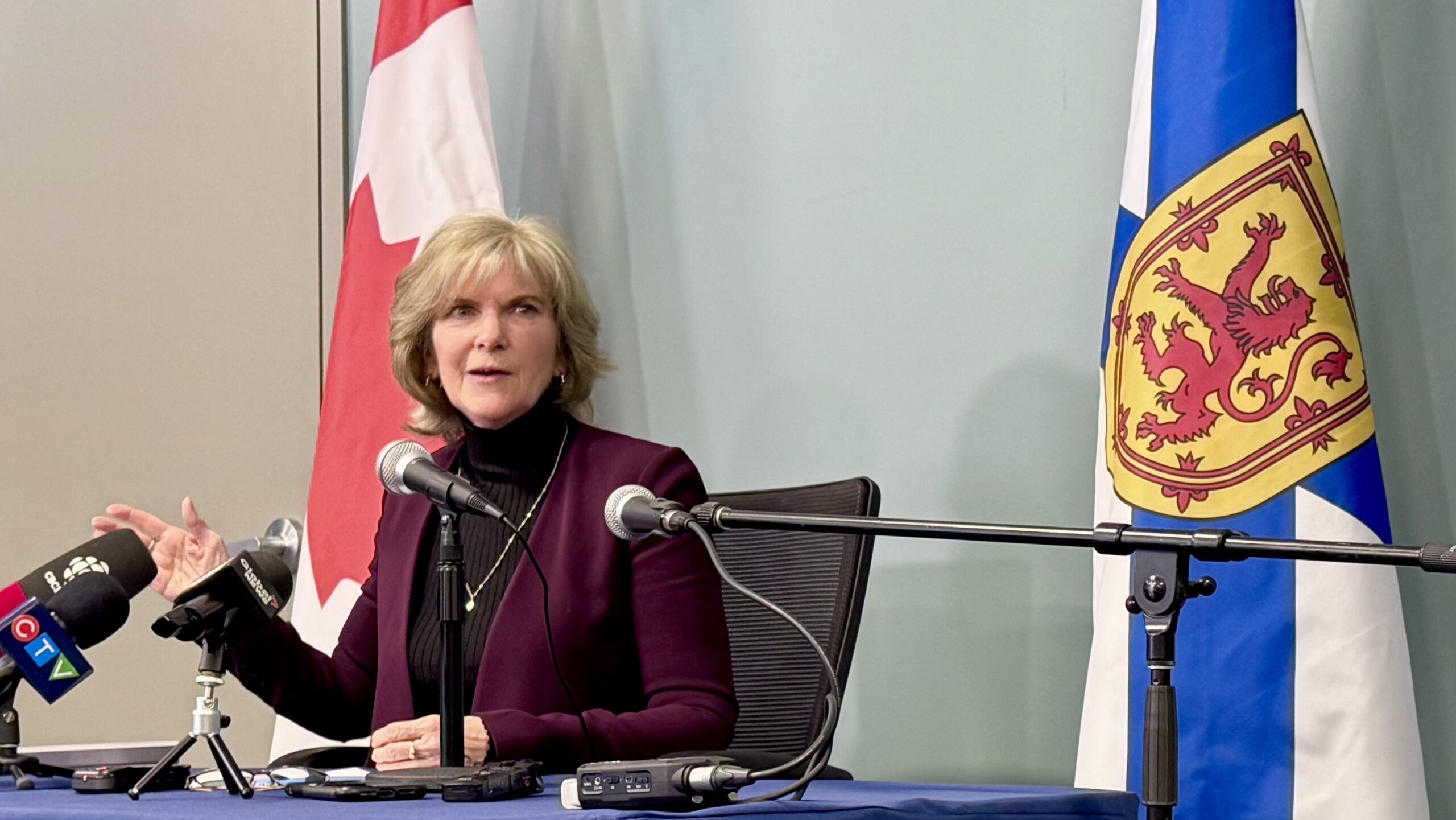 A woman wearing a red blazer sits at a desk with microphones in front of her