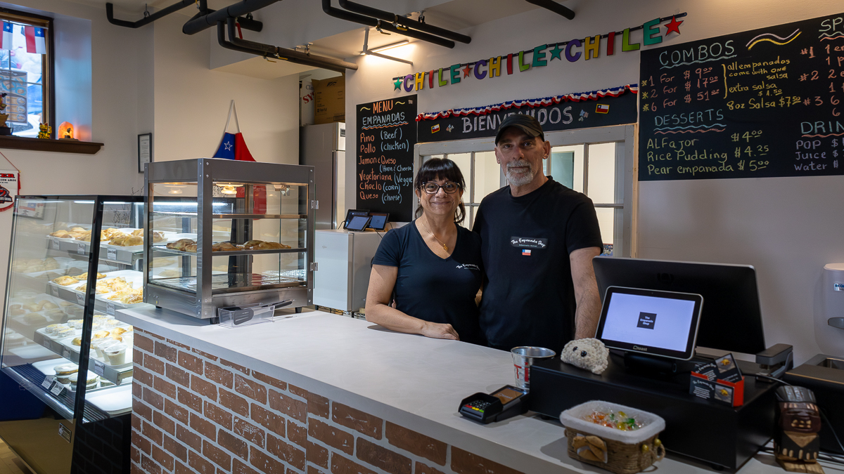 a man and women stand behind a brick counter with a white top. Behind them is a black chalkboards with a menu list of combos and desserts for empanadas. To the left of them is a glass display case full of empanadas, dinner pastries shaped like a half moon.