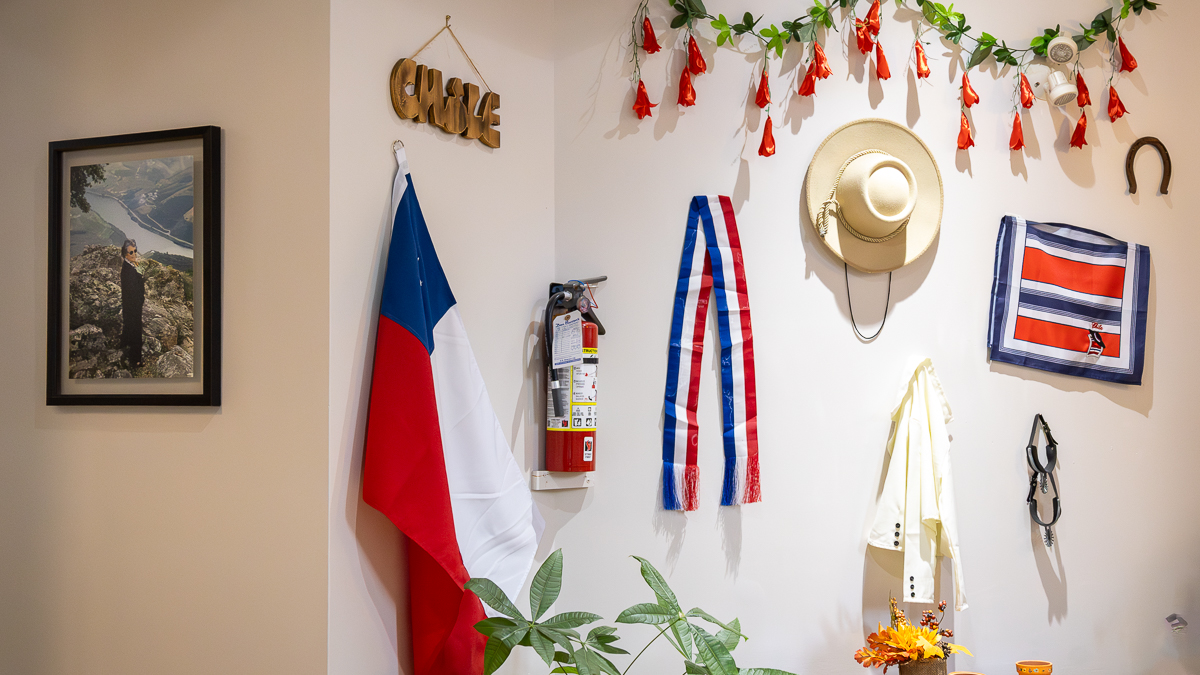 A white wall has multiple items from Chile hanging on it. On the left is a portrait of a woman. On the right is the flag of Chile, a vine of red flowers, a Chilean scarf, a beige hat, a white jacket and boot spurs, a blue, red and white cloth, and a horse shoe.