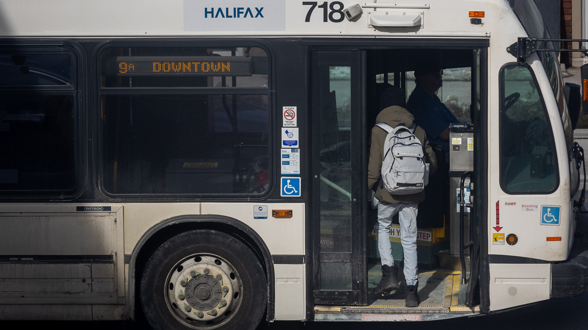 A man gets on a white Halifax transit bus. he is wearing a green jacket, jeans, and a grey backpack. The bus sign says its the 9A heading downtown.