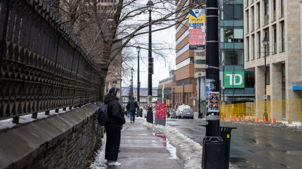 a woman waits at a bus stop on the sidewalk in downtown Halifax. she is wearing a black coat and pants with a black backpack. she stands along a stone and metal fence.